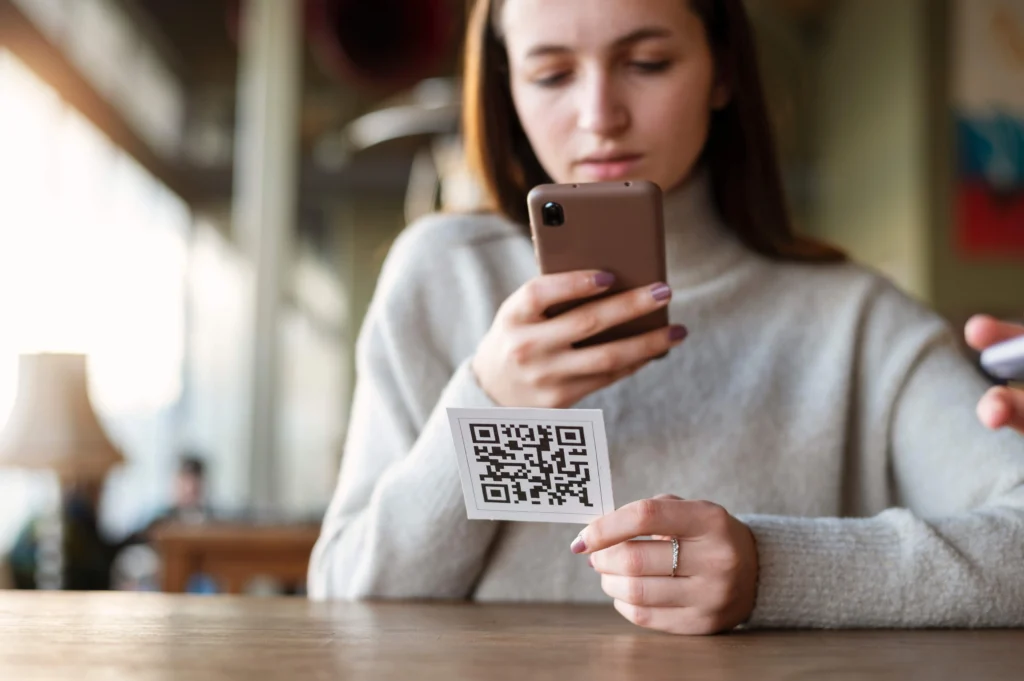 A woman holding a QR code card and scanning it with her smartphone, highlighting the potential risk of QR code scams in public places.